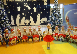 Dziewczynka recytuje wiersz, trzyma w ręku mikrofon w tle dzieci siedzą na krzesłach na tle dekoracji świątecznej, choinek.